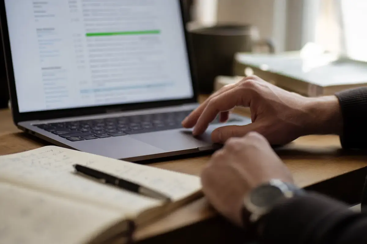Hands on a laptop that shows an interview transcript, with a notebook and pen next to it